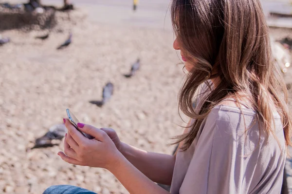Όμορφη γυναίκα χρησιμοποιώντας το κινητό της τηλέφωνο ενώ κάθεστε στον ξύλινο πάγκο. Casual στυλ - τζιν και πουκάμισο. Ευτυχισμένη γυναίκα χρησιμοποιώντας smartphone σε μια ηλιόλουστη ημέρα — Φωτογραφία Αρχείου