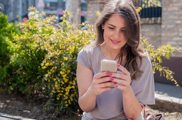 Νεαρή γυναίκα κάθεται σε ένα παγκάκι και χρήση κινητού τηλεφώνου. Attractivk με ένα πράσινο φόντο αόριστες. Χαρούμενη γυναίκα γραπτών μηνυμάτων με το smartphone σε ένα πάρκο — Φωτογραφία Αρχείου