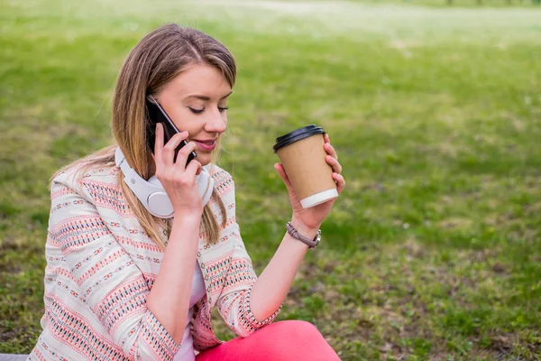 Mujer joven con café para ir en la mano usando un teléfono móvil y auriculares en el parque — Foto de Stock