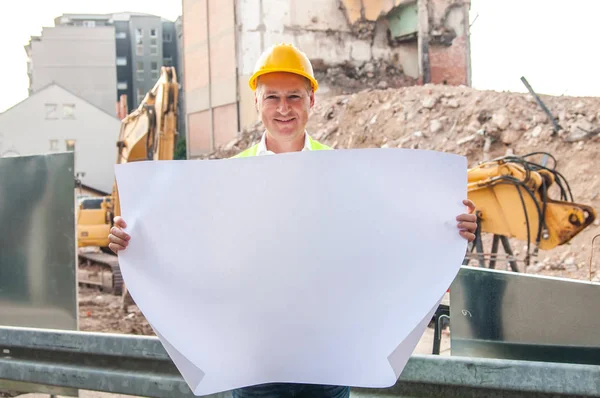 portrait of smiling civil engineer working with documents on construction site. Construction engineer in yellow hardhat inspecting blueprints on building site