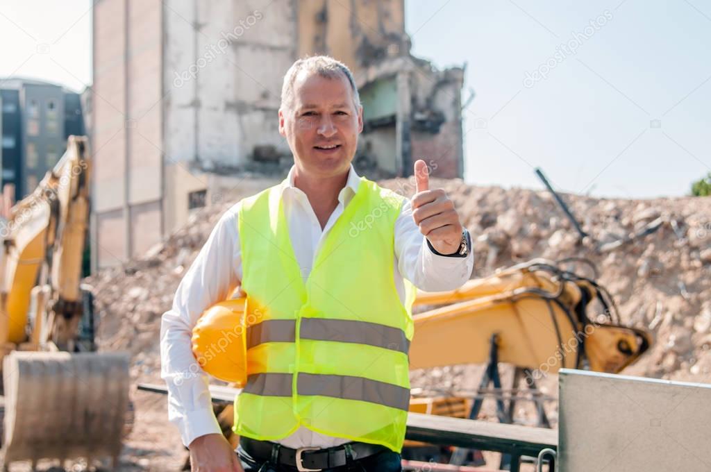 portrait of smiling civil engineer holding hardhat and showing thumb up gesture on construction site