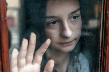 sad teenage girl looking out of window on cold autumn day. Lonely thoughtful girl touching glass clipart