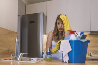 Lastik eldiven ve önlük sepeti ile temizlik malzemeleri mutfak önünde tutan sinir bozucu kadın. Mutsuz kadın temizlik büyük mutfak. genç çekici üzgün ve depresyonda kadın veya temizlik ev kadını. Yorgun ve çaresiz tembel ev ödevi ve kubbe