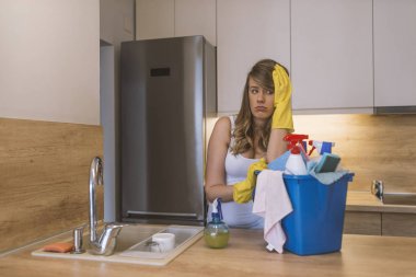 Lastik eldiven ve önlük sepeti ile temizlik malzemeleri mutfak önünde tutan sinir bozucu kadın. Mutsuz kadın temizlik büyük mutfak. genç çekici üzgün ve depresyonda kadın veya temizlik ev kadını. Yorgun ve çaresiz tembel ev ödevi ve kubbe