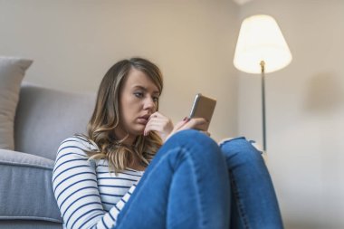 Kötü sms alma genç üzücü rahatsız kadın portresi. Önden Görünüm evde oturma odasında katta oturan telefon kontrol üzgün bir kadın. Üzücü bir haber. Genç kadın ile hareket eden telefon mesajı okur. Portre portre üzgün üzgün şüpheci BM