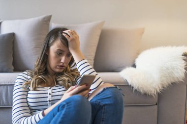Kötü sms alma genç üzücü rahatsız kadın portresi. Önden Görünüm evde oturma odasında katta oturan telefon kontrol üzgün bir kadın. Üzücü bir haber. Genç kadın ile hareket eden telefon mesajı okur. Portre portre üzgün üzgün şüpheci BM