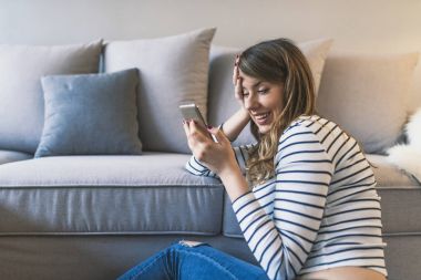 Onu bi güzel mesaj. Seyir onun akıllı telefon ve halı üzerinde evde otururken gülümseyen çekici genç kadın. Gülümseyen ev rahatlatıcı adlı genç kadın, o bir smartphone ve manifatura, teknoloji ve iletişim kavramı kullanıyor. Genç hipst