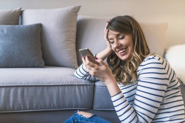 Onu bi güzel mesaj. Seyir onun akıllı telefon ve halı üzerinde evde otururken gülümseyen çekici genç kadın. Gülümseyen ev rahatlatıcı adlı genç kadın, o bir smartphone ve manifatura, teknoloji ve iletişim kavramı kullanıyor. Genç hipst