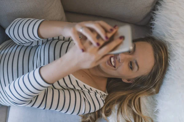 Lächelnde junge Frau zu Hause, die es sich auf der Couch gemütlich macht, benutzt sie — Stockfoto