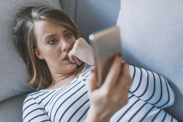 Kötü sms alma genç üzücü rahatsız kadın portresi. Önden Görünüm evde oturma odasında katta oturan telefon kontrol üzgün bir kadın. Üzücü bir haber. Genç kadın ile hareket eden telefon