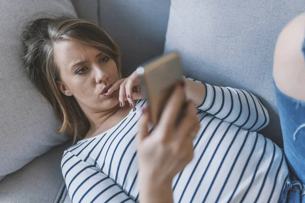 Portrait of young sad annoyed woman, receiving bad sms. Front view of a sad woman checking phone sitting on the floor in the living room at home. Sad news. Upset young woman with mobile phone