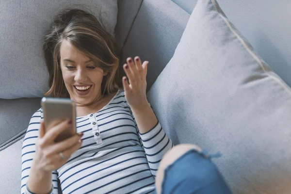 The message that brightened her day. Some status updates just make you smile. Casual shocked young woman looking at cellphone on sofa at home. Adorable girl smiling and reading message on phone