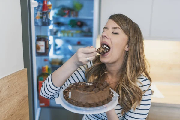 Close-up of young woman standing in front of open refrigerator eating cake. Woman in front of the fridge with cake on dessert plate in hand. Woman Eating Unhealthy Chocolate Cake in Front of Open Refrigerator