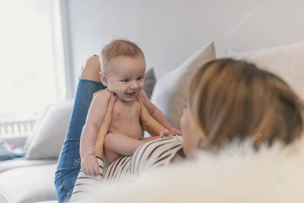 Mother and child on a bed. Mom and baby boy in diaper playing in sunny living room. Parent and little kid relaxing at home. Family having fun together. Bedding and textile for infant nursery. Portrait of beautiful mom playing with her 4 months old ba