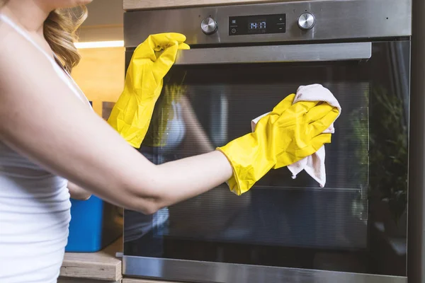 Close up of female hand with yellow protective gloves cleaning oven door. young smiling woman in protective glove with rag cleaning oven. Girl polishing kitchen. People, housework, housekeeping, cleaning concept