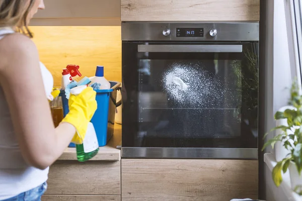 Close up of female hand with yellow protective gloves spraying detergent from bottle on the oven door.  young smiling woman in protective glove with rag cleaning oven. Girl polishing kitchen. People, housework, housekeeping, cleaning concept