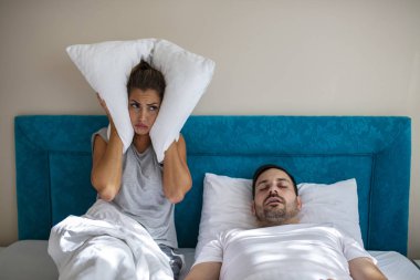 Genç çiftlerin horlamalarıyla ilgili problemleri var. Heteroseksüel çift yatakta, erkek uyuyor ve ağzı açık horluyor, yorgun bir kadın ise kafasında yastıkla yatakta horlamaktan rahatsız oluyor..