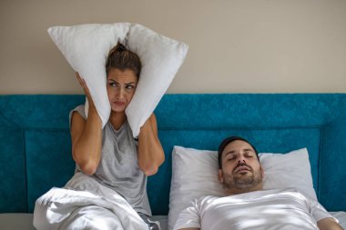 Young couple have problem with man's snoring. Heterosexual couple in bed, man sleeps and snoring with mouth open, while a tired woman irritated by snoring sitting on bed with a pillow on her head. clipart