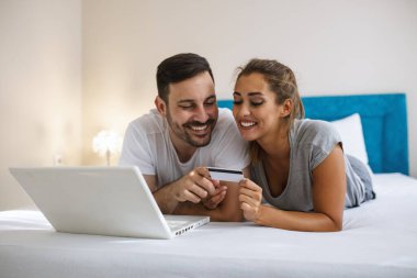 Beautiful young amorous couple using laptop, on bed. Happy young couple shopping online while lying on their bed. Young couple surfing on internet and shopping with laptop on bed. clipart