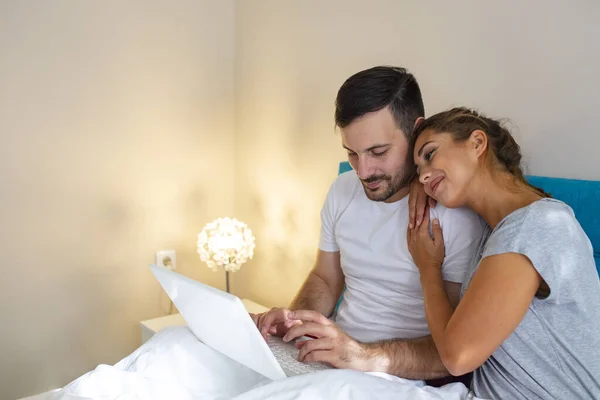 Side view of young couple with digital laptop with blank screen resting on bed at home. Young married couple lying on bed at home and using laptop. Attractive woman embracing husband while using a laptop in bed