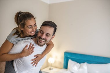 Young playful couple having fun while piggybacking. Handsome man in white T-shirt carrying young attractive woman on shoulders while spending free time at home