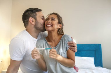 Young couple is happy because of positive pregnancy test. Affectionate couple finding out results of a pregnancy test in their bedroom. Family, parenting and medical concept clipart