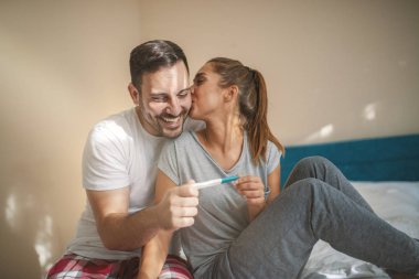 Happy couple with positive pregnancy test. Happy excited couple making positive pregnancy test and celebrating. Cheerful couple finding out results of a pregnancy test sitting on bed clipart