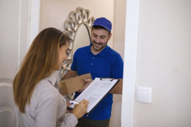 Evdeki kuryeden paket için imza atan kadın. Gülümseyen teslimatçı elinde karton bir kutu tutarken güzel genç bir kadın panoya imza atıyor.