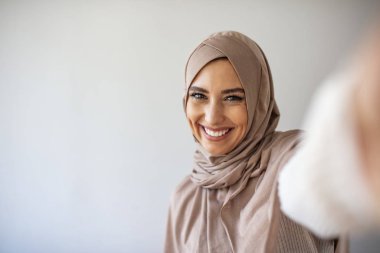 Arap güzel bir kadın gülümsüyor ve gri arka planda cep telefonuyla fotoğraf çekiyor. Genç Müslüman kadının portresi cep telefonuyla selfie çekerken poz veriyor.