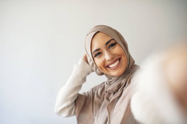 Arap güzel bir kadın gülümsüyor ve gri arka planda cep telefonuyla fotoğraf çekiyor. Genç Müslüman kadının portresi cep telefonuyla selfie çekerken poz veriyor.