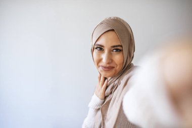 Arap güzel bir kadın gülümsüyor ve gri arka planda cep telefonuyla fotoğraf çekiyor. Genç Müslüman kadının portresi cep telefonuyla selfie çekerken poz veriyor.