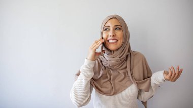 Gri arka planda cep telefonuyla konuşan 20 'li yaşlarda, İslami başörtülü genç bir Arap kadının portresi. Cep telefonu kullanan mutlu Müslüman kadının portresi