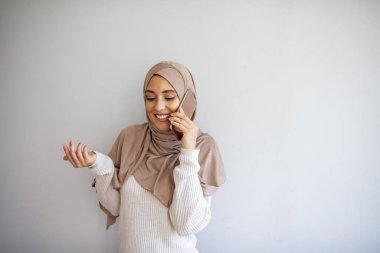 Cep telefonu kullanan tesettürlü Asyalı kadın. Akıllı telefon kullanan tesettürlü Müslüman bir kız. Gri arka planda cep telefonu tutan gülümseyen genç bir Arap kadının portresi