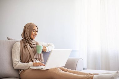 Evde tesettürlü mutlu Müslüman kadın. Kahve içip internetten alışveriş yapan bir kız, fotokopi odası. Genç bir iş kadınının dizüstü bilgisayar kullanırken ve evde kahve içerken fotoğrafı.. 