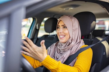 Araba süren ve bağıran Müslüman bir kadın. Negatif insani duygular ifade edilebilir. Kızgın kadın sürücü profili. Kızgın kadın araba kullanıyor. Hoşnutsuzluk ifadesi olan kız aktif olarak arabanın direksiyonunda el kol hareketi yapıyor..