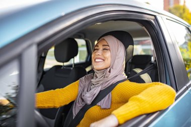 Başörtülü genç Asyalı Müslüman kadın arabanın içinde gülümsüyor. Güzel, beyaz, Müslüman bir kadın, bir araba çekiminde hafif tesettüre bürünmüş. Güzel Müslüman kadın, dişlek gülüşlü araba kullanıyor.. 