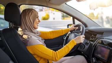 Güzel, beyaz, Müslüman bir kadın, bir araba çekiminde hafif tesettüre bürünmüş. Güzel Müslüman kadın, dişlek gülüşlü araba kullanıyor. Orta Doğulu araba kullanan bir kadının portresi, modern bej bir Abaya giyiyor..