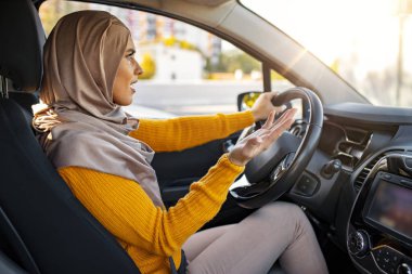 Müslüman Kadın arabasını çok agresif kullanıyor ve eliyle el hareketi yapıyor. Yakın plan portre kızgın, agresif Müslüman kadın araba kullanıyor, birine bağırıyor, elleri havada trafik geçmişinde.