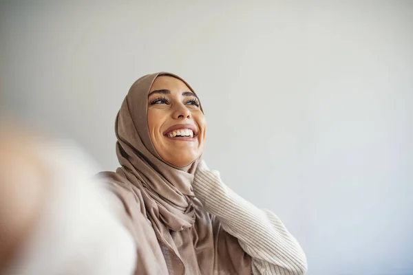 幸せな若いイスラム教徒の女性は自宅で携帯電話で自画像を取ります 携帯電話でヒジャーブの幸せなアラブ女性が自撮りをしています 笑顔の少女の肖像 ポーズ — ストック写真