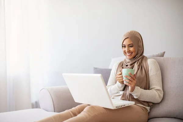 Young Arabic female entrepreneur wearing a hijab working on a laptop and drinking coffee while sitting at living room. Muslim woman with laptop at home.  Attractive young woman in head scraf using laptop while sitting on couch