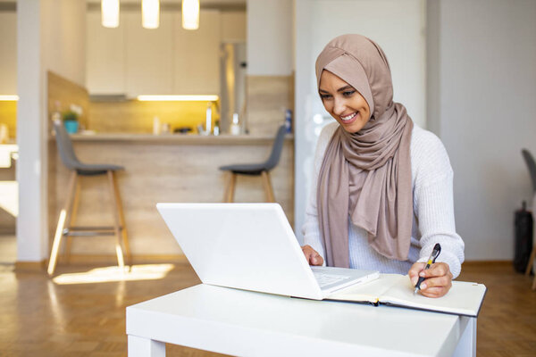 Pretty woman wearing hijab in front of laptop search and doing office work with different face expression isolated in home background - office, business, finance and work station concept