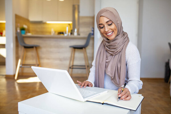 Young saudi business woman working in at home, pointing on laptop, copy space. Muslim female working with computer in the room , writing paper. Young muslim woman using laptop