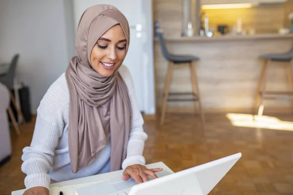 美しい女性は イスラムのスカーフを着て ノートに鉛筆の書き込みを保持木製のテーブルの上に座って コンピュータでかわいい笑顔 ラップトップコンピュータで働く魅力的な女性アラビア語と机の上の書類 — ストック写真