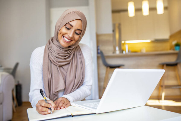 Muslim woman working in home office. Arabian woman dressed in religious veil is working on her laptop, looking at screen and typing on keyboard. She seems to be concentrated and glad using various softwares and online services. 