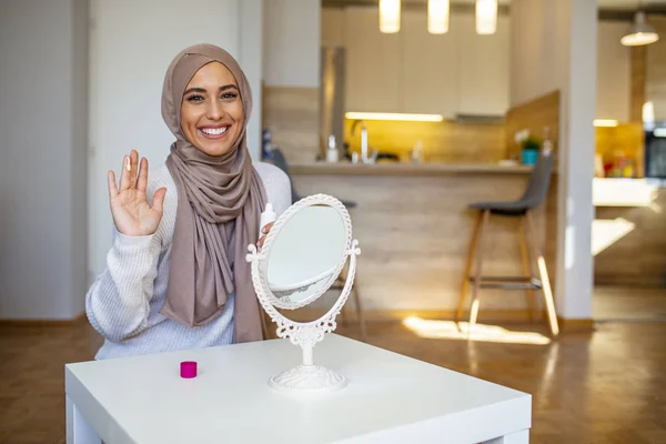 Smiling muslim lady applying moisturizing face cream, skin care, cosmetology. Muslim woman spreading cream over her face while looking in the mirror. Beauty treatment.