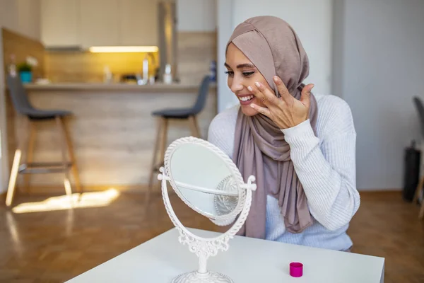 Muslim woman putting on moisturizer on her facial. Smiling young Muslim woman applying cream to face and looking to mirror. Muslim woman putting anti-aging cream on her face