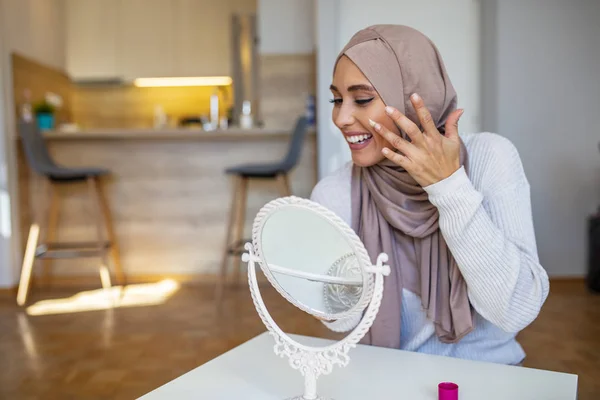 手鏡を見て顔クリームを塗るムスリム女性の笑顔 家で鏡を見て顔にクリームを塗る若いイスラム教徒の女性 クリームを入れてみましょう — ストック写真