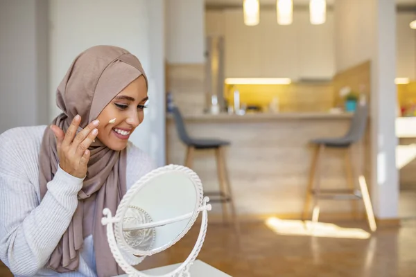 Muslim woman spreading cream over her face while looking in the mirror. Beauty treatment. Female putting on moisturizer on her facial. Smiling Muslim woman applying cream to face and looking to mirror