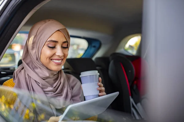 高級車の後部座席に座っているヒジャーブを持つ美しいイスラム教徒のアラビア人女性 彼女はタブレットコンピュータを見に行くためにコーヒーを飲む 幸せな美しいです若いですムスリム女性の肖像車の後部座席に座って — ストック写真