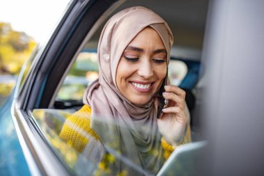 Genç Müslüman iş kadını arabanın arka koltuğunda telefonla konuşuyor ve elinde dijital bir tablet tutuyor. Arabadaki güzel Müslüman iş kadını.